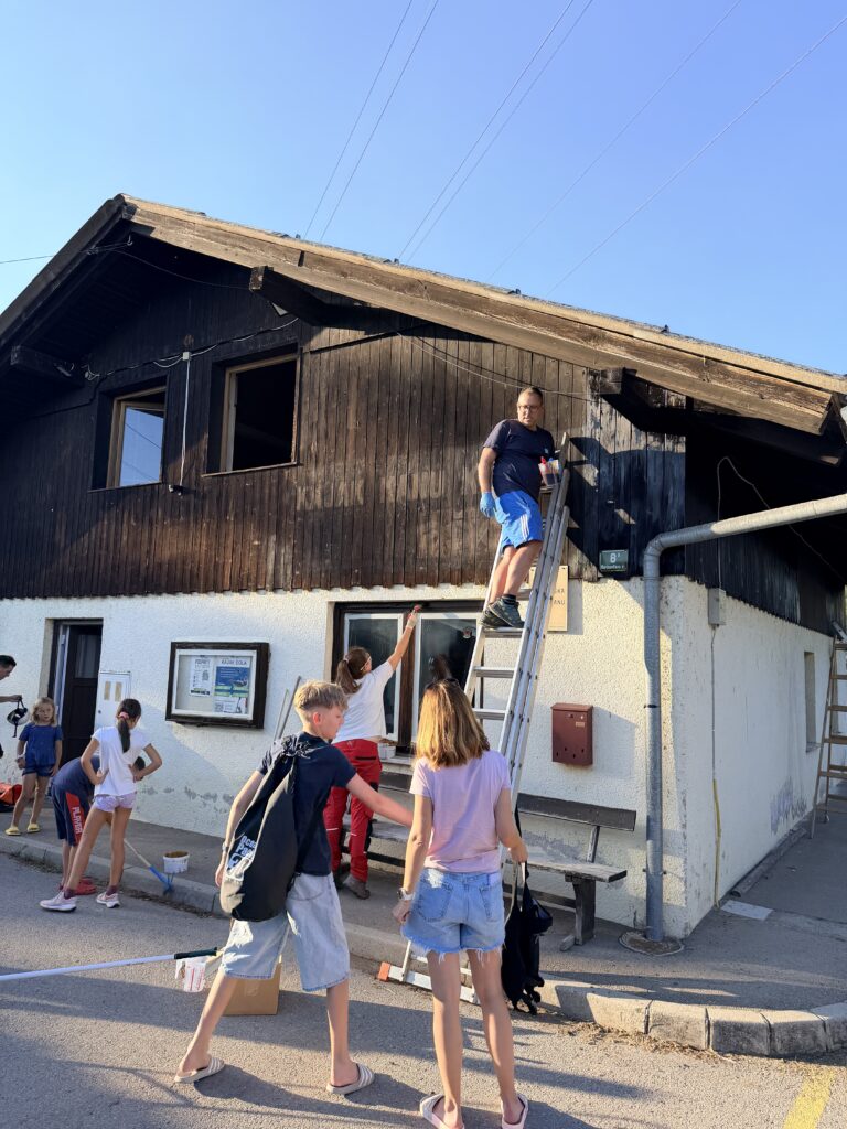 Več generacij članov pri barvanju fasade klubskega doma, z moškim na lestvi in sodelujočimi ob vhodu.