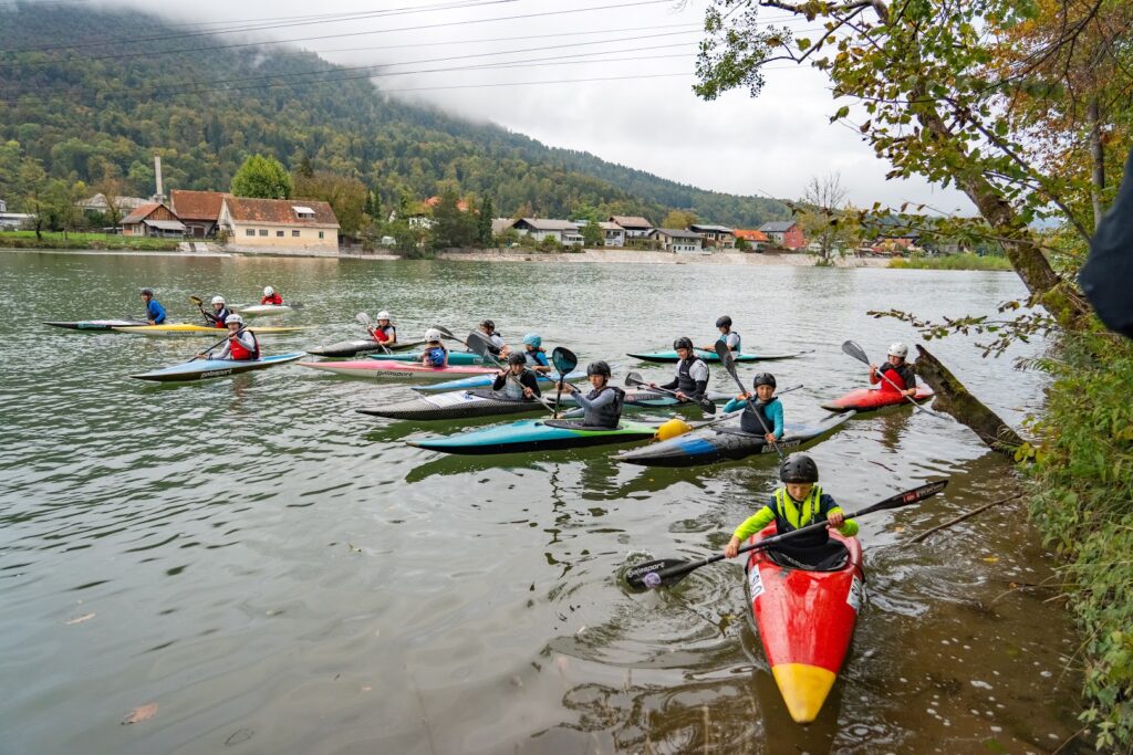 Široki kader skupine kajakašev ob bregu Save; v ospredju rdeč čoln, zadaj Šmarna gora.