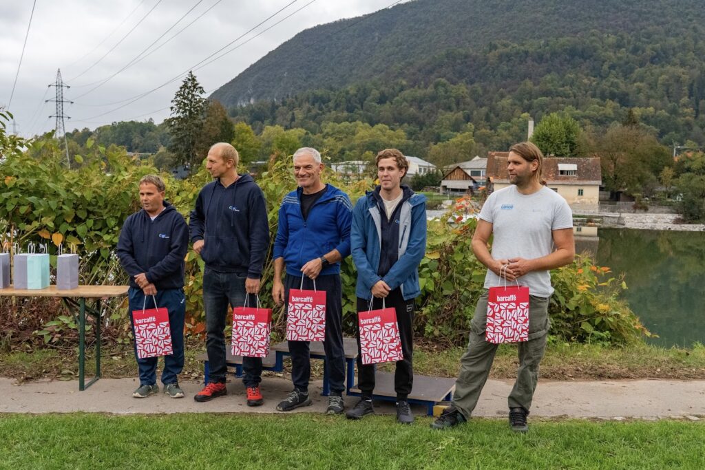 Pet trenerjev z rdečimi darilnimi vrečkami ob Savi, s Šmarno goro v ozadju.